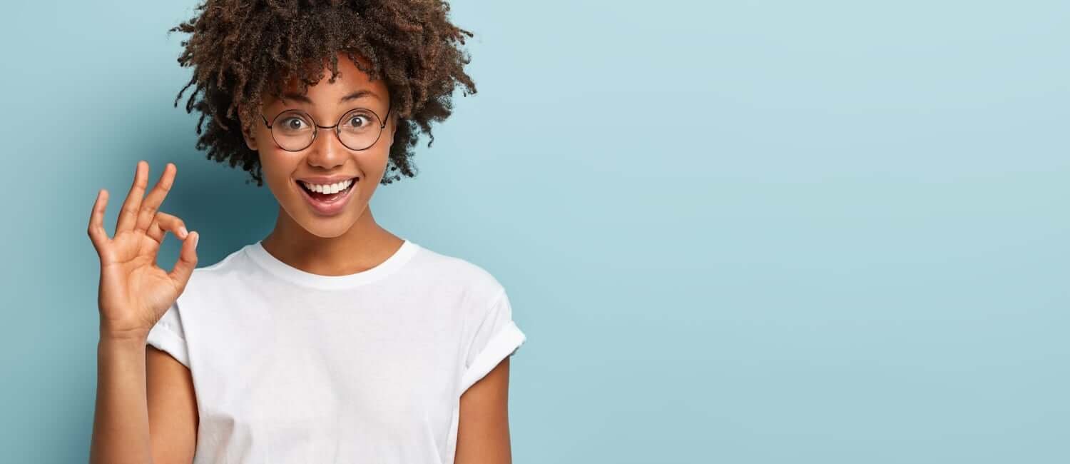 Horizontal shot of cheerful Afro American woman makes okay gesture, agrees with good proposal, wears spectacles and white casual t shirt, says incredible offer, isolated over blue background La medit de la semaine